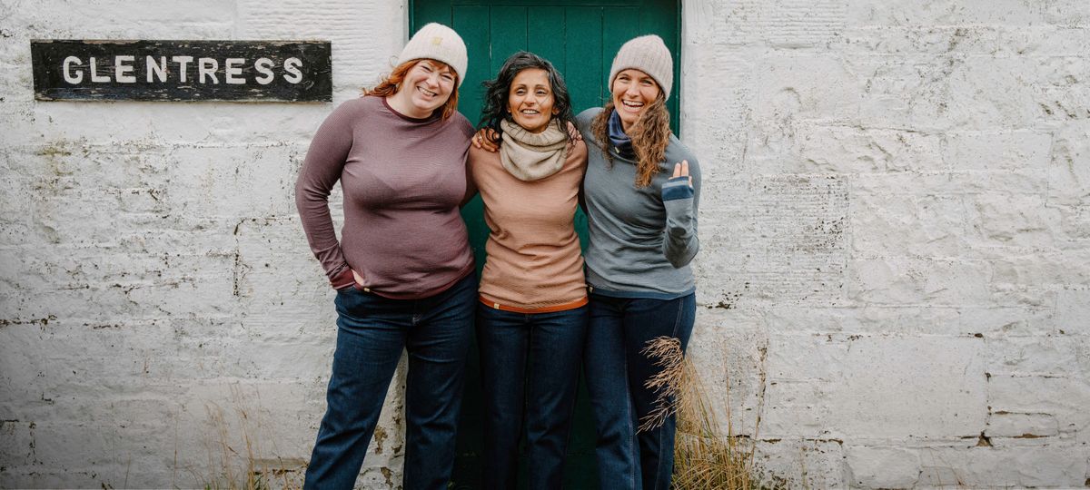 Three women standing together in front of a white textured wall with a green door and a black sign reading 'GLENTRESS'. They are wearing knit beanies and layered tops, and the scene appears to be a styled outdoor or rustic storefront setting.