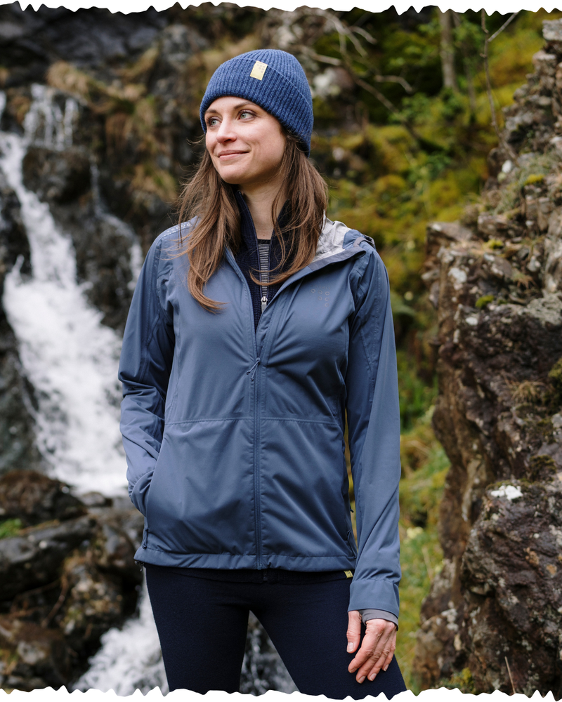 Woman wearing a blue jacket and beanie standing in a natural setting with waterfalls and rocks.