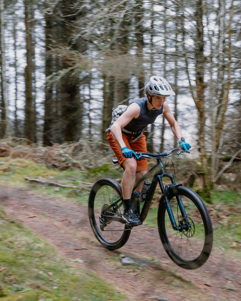 A mountain biker wearing a gray helmet, black sleeveless top, orange shorts, and blue gloves rides a black and blue bike on a dirt trail through a forest.