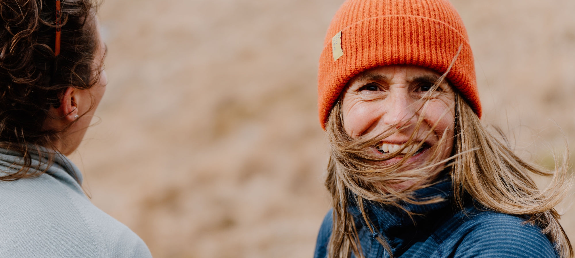 A woman outdoors wearing a bright orange knit beanie and a blue jacket, smiling while windblown hair blows across her face. Another person is visible from the side in the foreground, with a soft sandy background.
