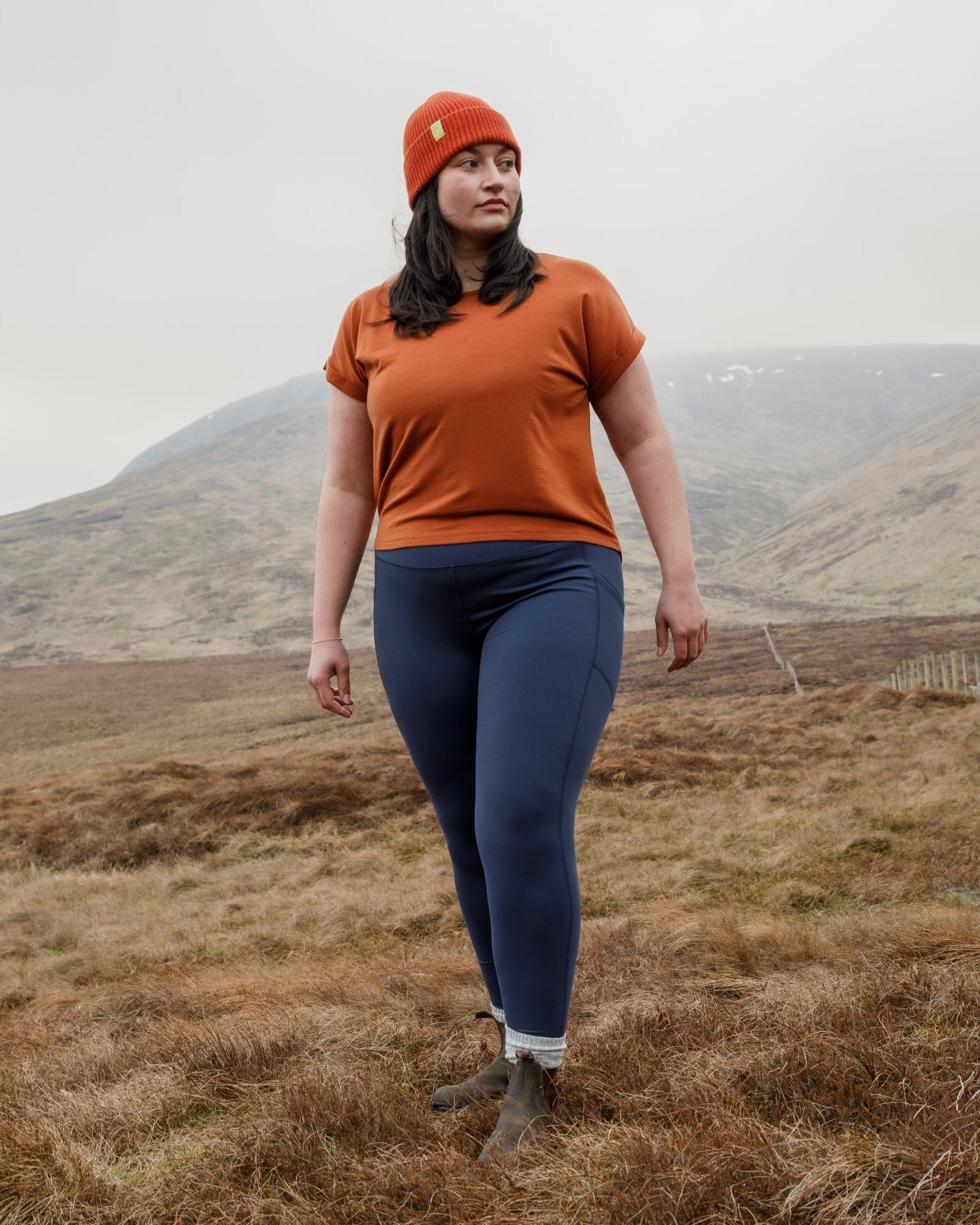 Person wearing an orange shirt and blue leggings standing in a field with mountains in the background