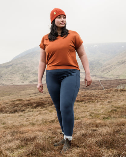 Person wearing an orange shirt and blue leggings standing in a mountainous landscape.