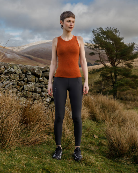 Woman wearing a FINDRA orange Merino tank top and black leggings standing in a natural landscape with mountains and trees.
