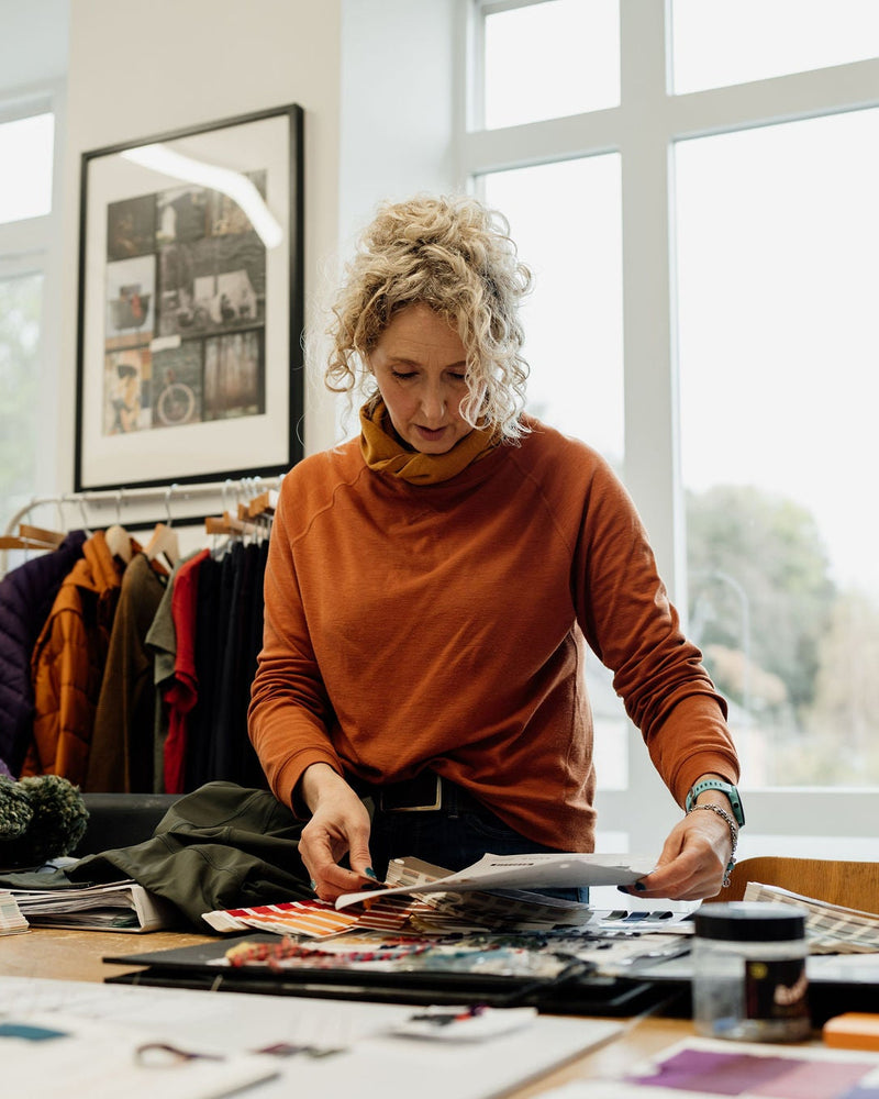 Alex Feechan - founder and CEO of FINDRA in an orange sweater working at a desk with design materials in a bright room.
