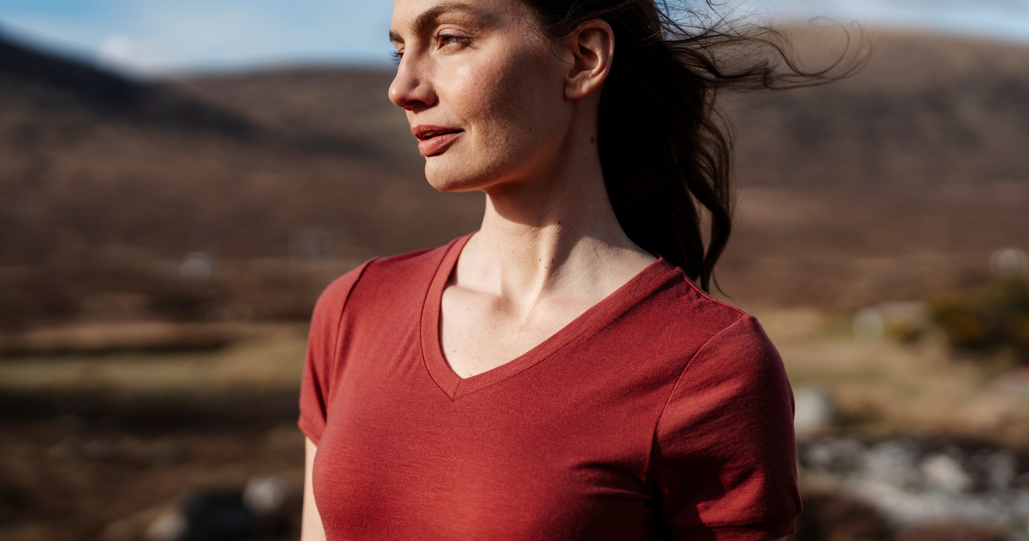 Woman in a red shirt standing in a scenic outdoor setting with mountains and a river.