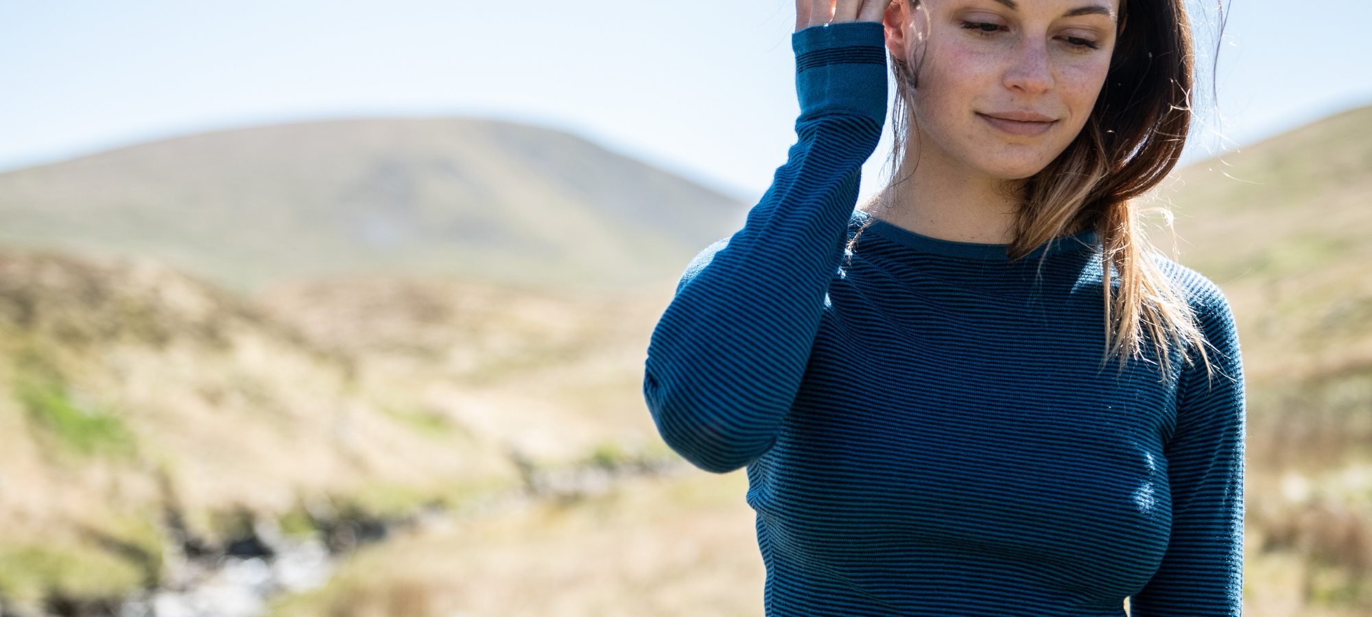 A woman wearing a dark teal ribbed knit long-sleeve top stands outdoors in a grassy, hilly landscape with her hand near her face.