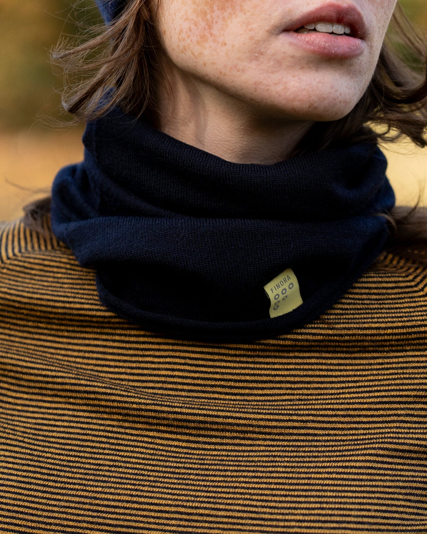 Woman wearing a neck warmer in dark navy, a navy beanie hat and a base layer in mustard and dark navy stripe in the outdoors in autumn
