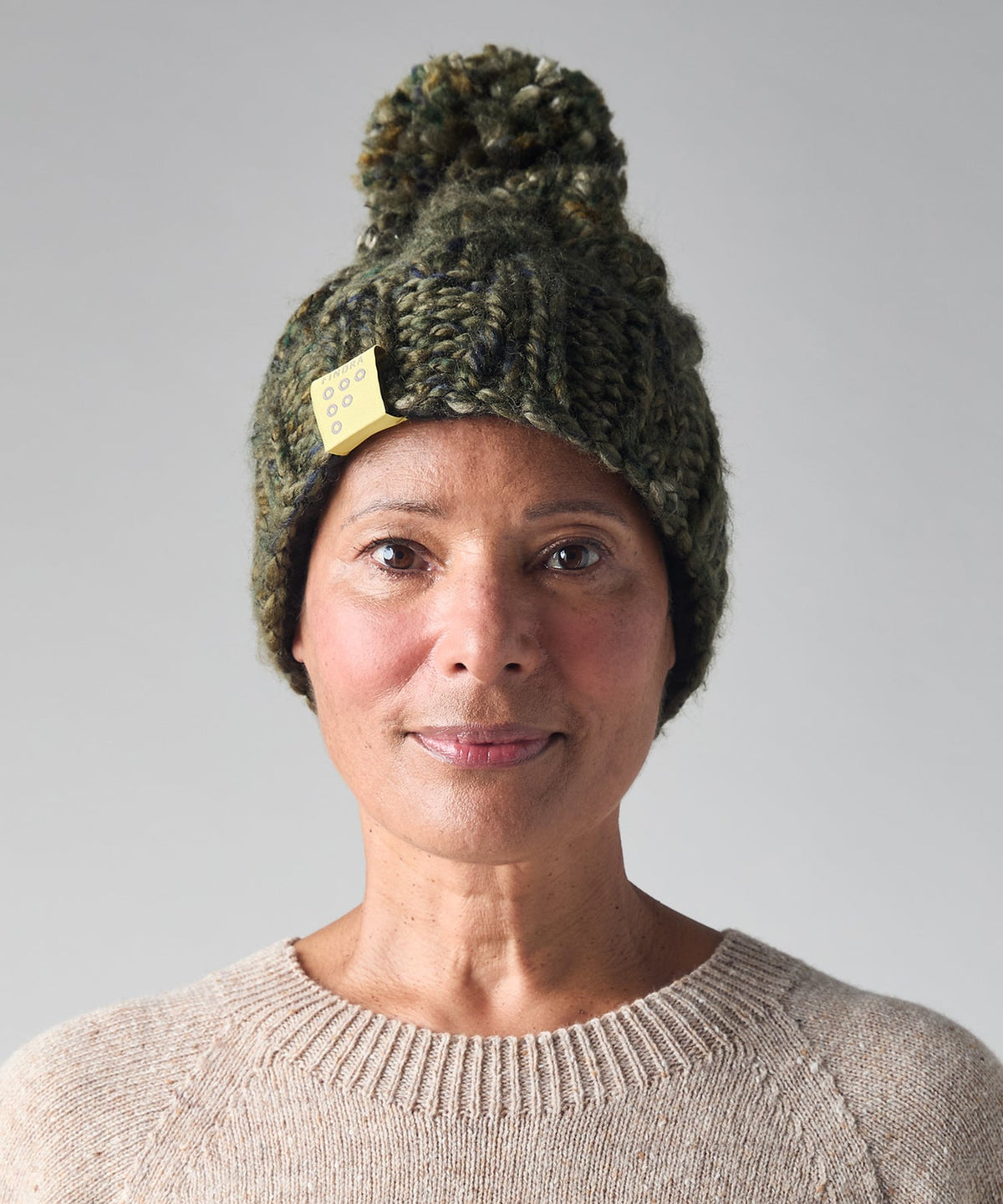 A woman wearing a dark green chunky knit beanie with a pom-pom and a small yellow tag, paired with a beige crewneck sweater, against a solid gray background.