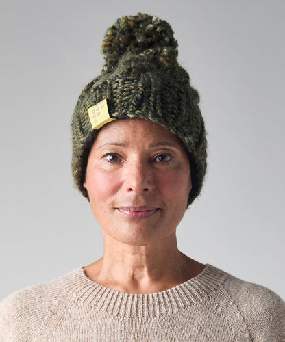 A woman wearing a dark green chunky knit beanie with a pom-pom and a small yellow tag, paired with a beige crewneck sweater, against a solid gray background.