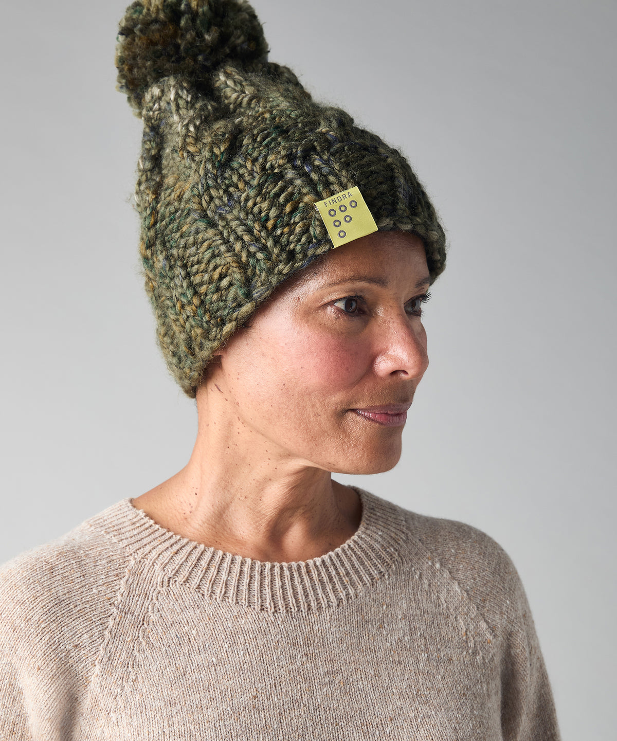 A woman wearing a chunky knit pom-pom beanie in a heathered olive green and brown colorway, paired with a beige knit crewneck sweater, photographed against a neutral gray background.