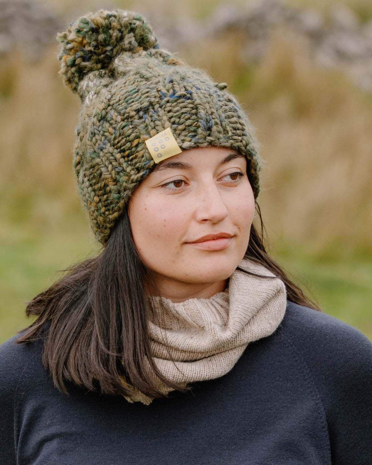 Woman in the outdoors wearing a chunky knotted Bobble hat in Green with a yellow label for FINDRA