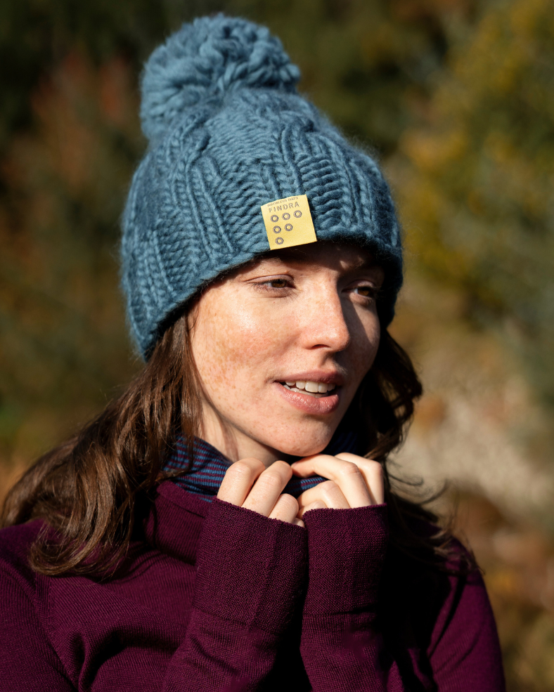Woman wearing a blue FINDRA  knitted Bobble Hat with a yellow tag in the outdoors in Autumn