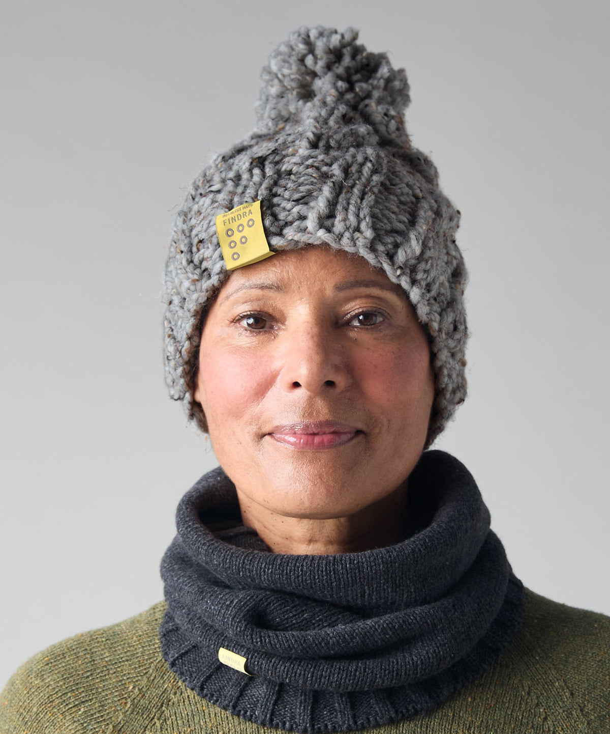 A woman wearing a chunky gray knit pom-pom beanie with a small yellow tag and a dark charcoal gray ribbed cowl neck warmer, paired with an olive green sweater, against a neutral gray background.