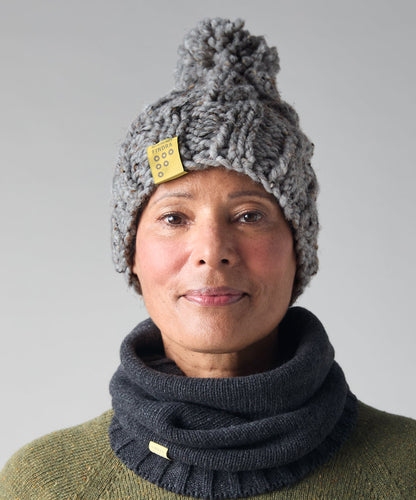 A woman wearing a chunky gray knit pom-pom beanie with a small yellow tag and a dark charcoal gray ribbed cowl neck warmer, paired with an olive green sweater, against a neutral gray background.
