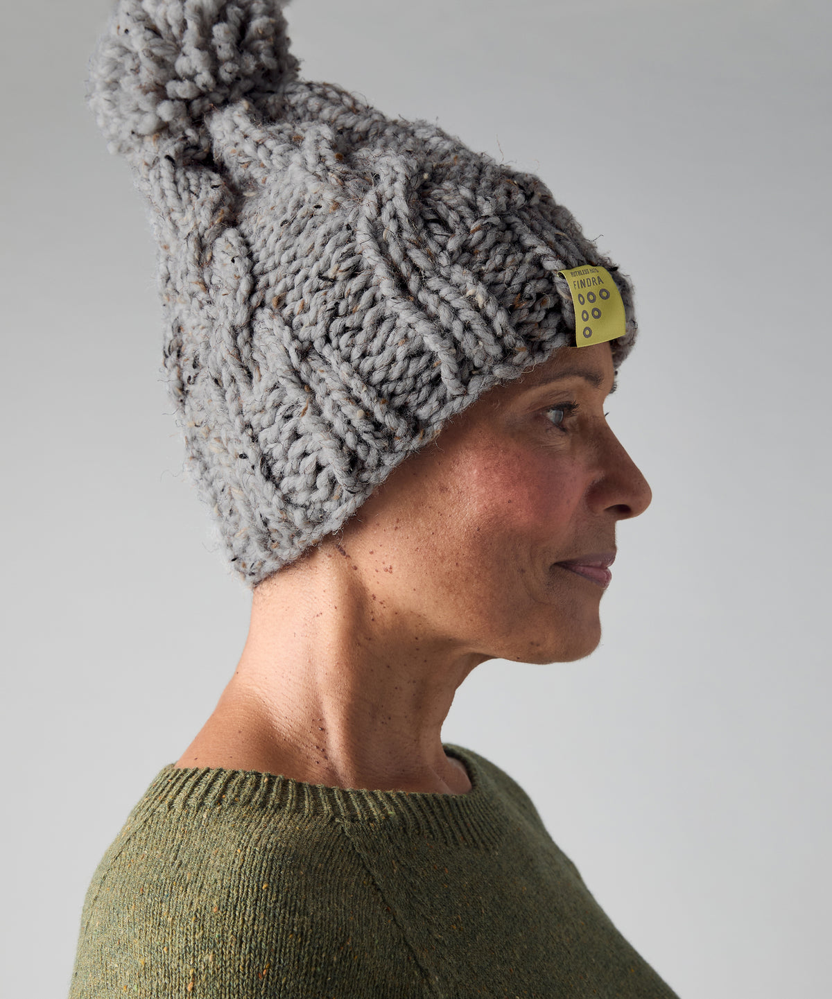 A side-profile portrait of a person wearing a chunky gray cable-knit beanie with a large pom-pom and a small yellow brand tag. The person is also wearing an olive green knit sweater against a plain light gray studio background.