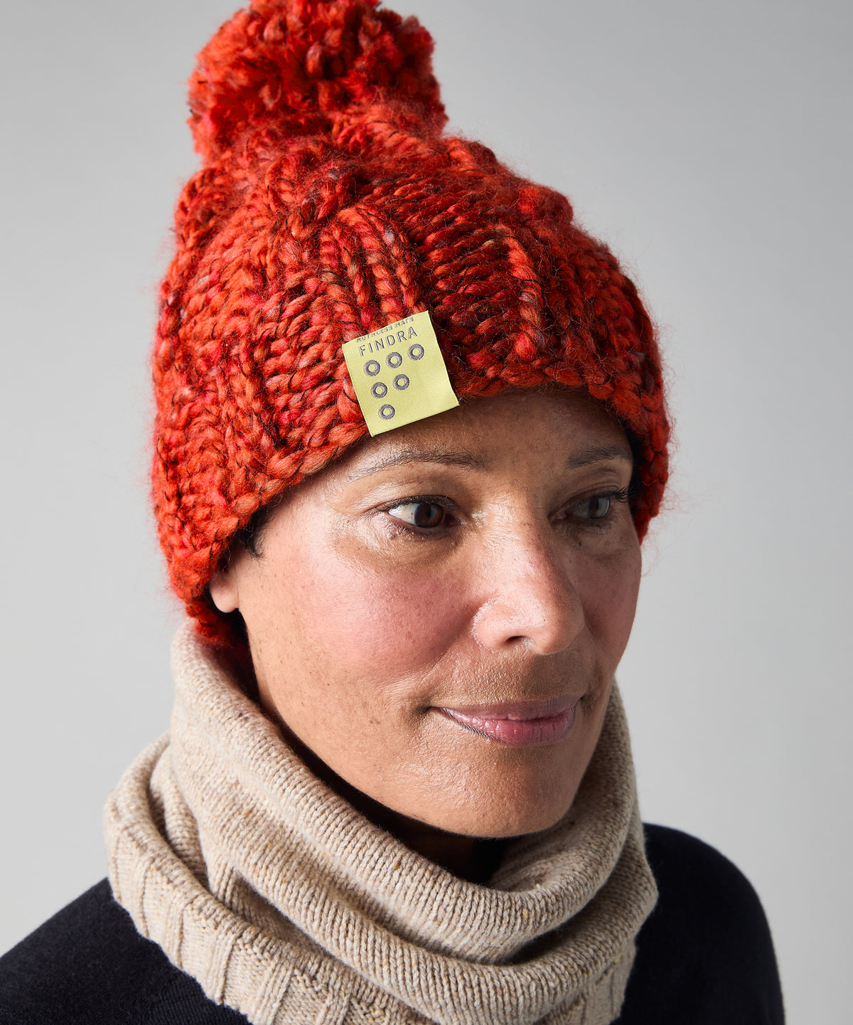 A close-up of a woman wearing a chunky knit red-orange bobble hat with a yellow brand patch and a beige knit cowl neck warmer, against a neutral gray background.