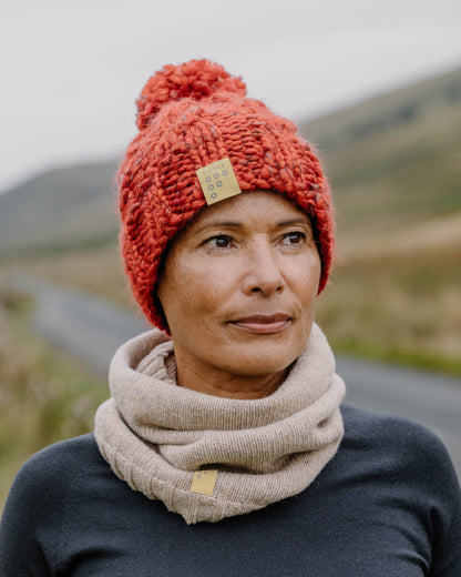 A woman outdoors wearing a chunky knit orange-red beanie with a pom-pom and a beige knit cowl scarf, both with small square brand patches. The background shows a blurred rural landscape.