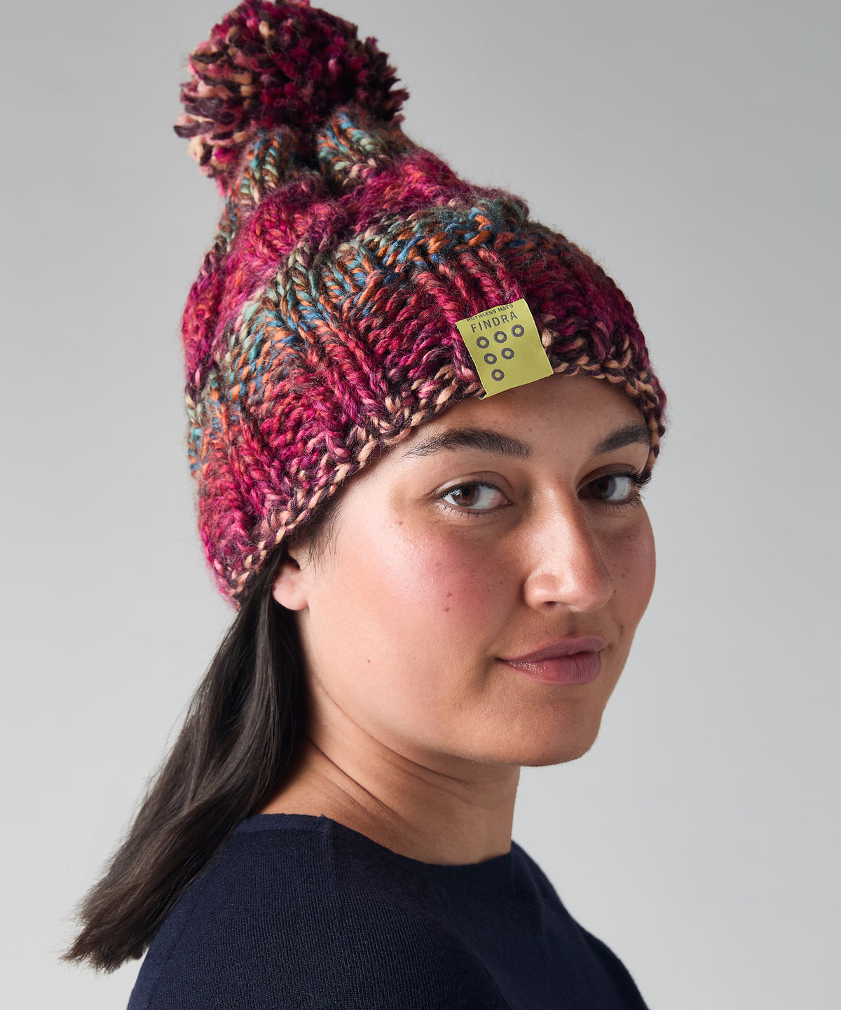 A woman wearing a chunky knit pom-pom beanie in a heathered mix of burgundy, pink, and teal, with a small yellow brand tag on the side.