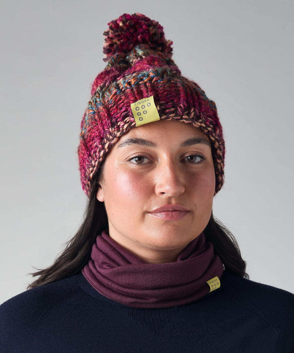 A woman wearing a dark navy long-sleeve top, a burgundy knit neck warmer, and a matching burgundy and brown knit beanie with a pom-pom. Small yellow tags are visible on both the beanie and the neck warmer, set against a plain light gray studio background.