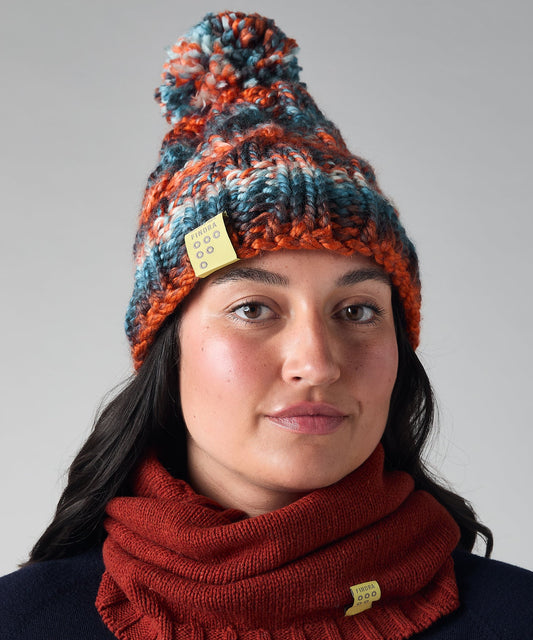 A woman wearing a chunky knit bobble hat and matching cowl neck warmer. The beanie features a marled pattern of orange, blue, and black yarn with a small yellow tag, and the neck warmer is a rust red color with a matching yellow tag.