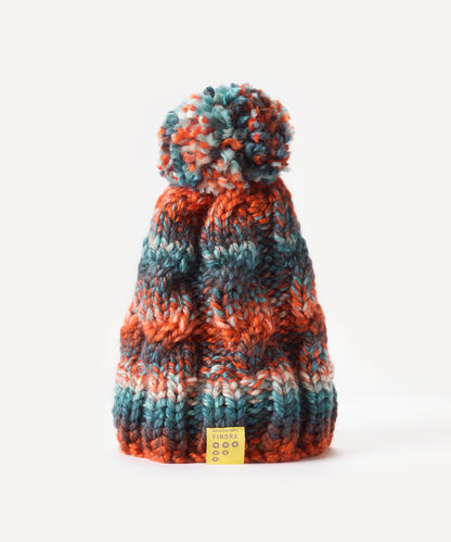A chunky knit bobble hat with a matching pom-pom, featuring horizontal stripes of rust orange, teal blue, and cream. A small yellow brand tag is attached at the bottom, set against a clean white background.