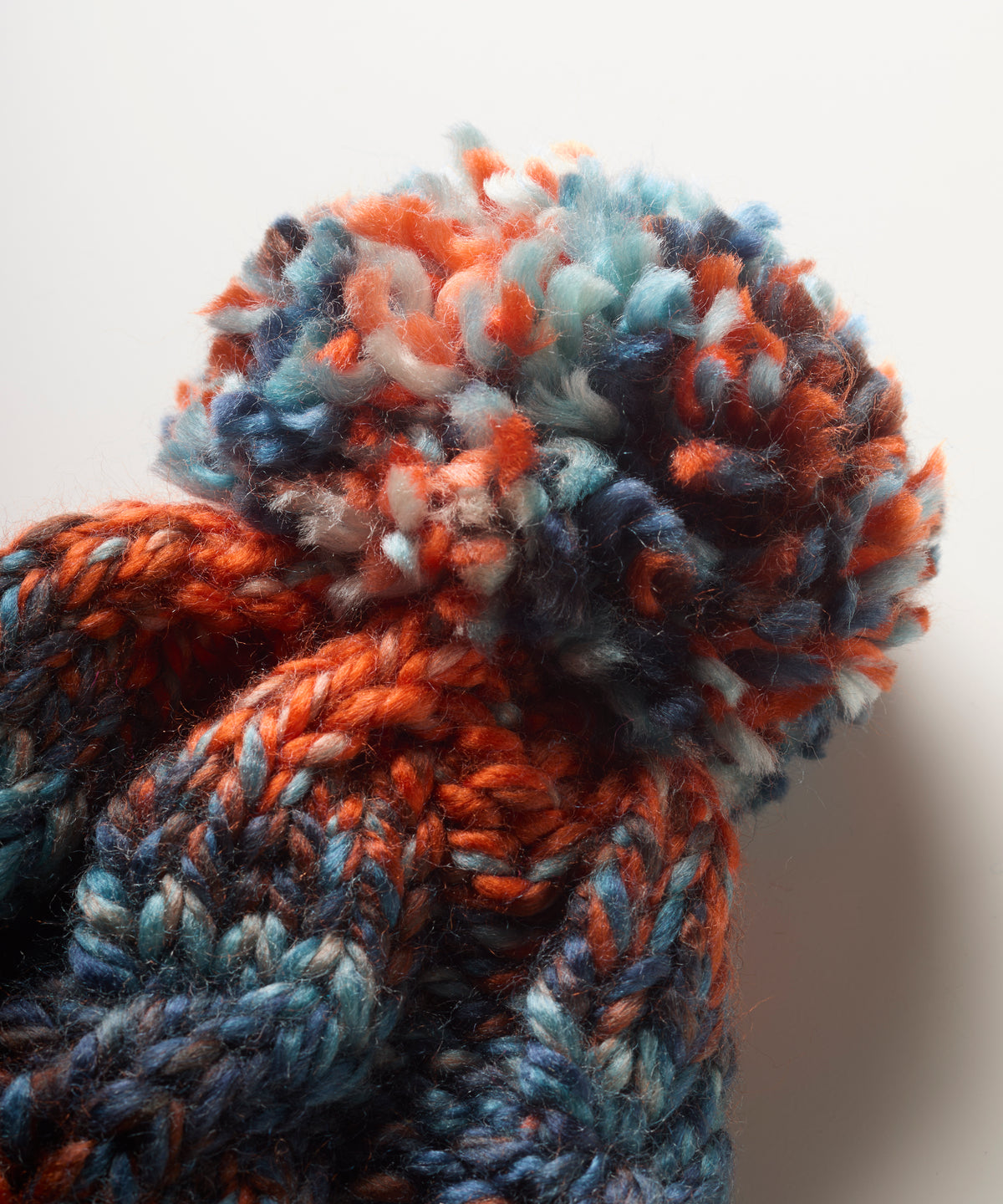 Close-up of a chunky knit bobble hat with a large fluffy pom-pom, made from variegued yarn in orange, blue, navy, and white on a plain light background.