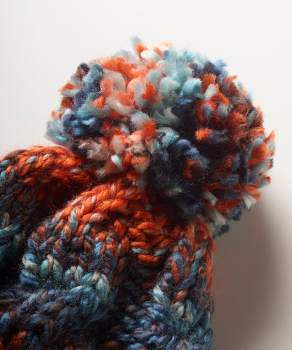Close-up of a chunky knit bobble hat with a large fluffy pom-pom, made from variegued yarn in orange, blue, navy, and white on a plain light background.