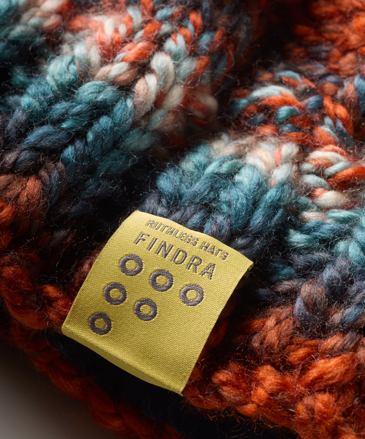 Close-up of a chunky knit hat in a variegated orange, teal blue, and cream yarn, featuring a yellow fabric brand label that reads 'FINDRA'.