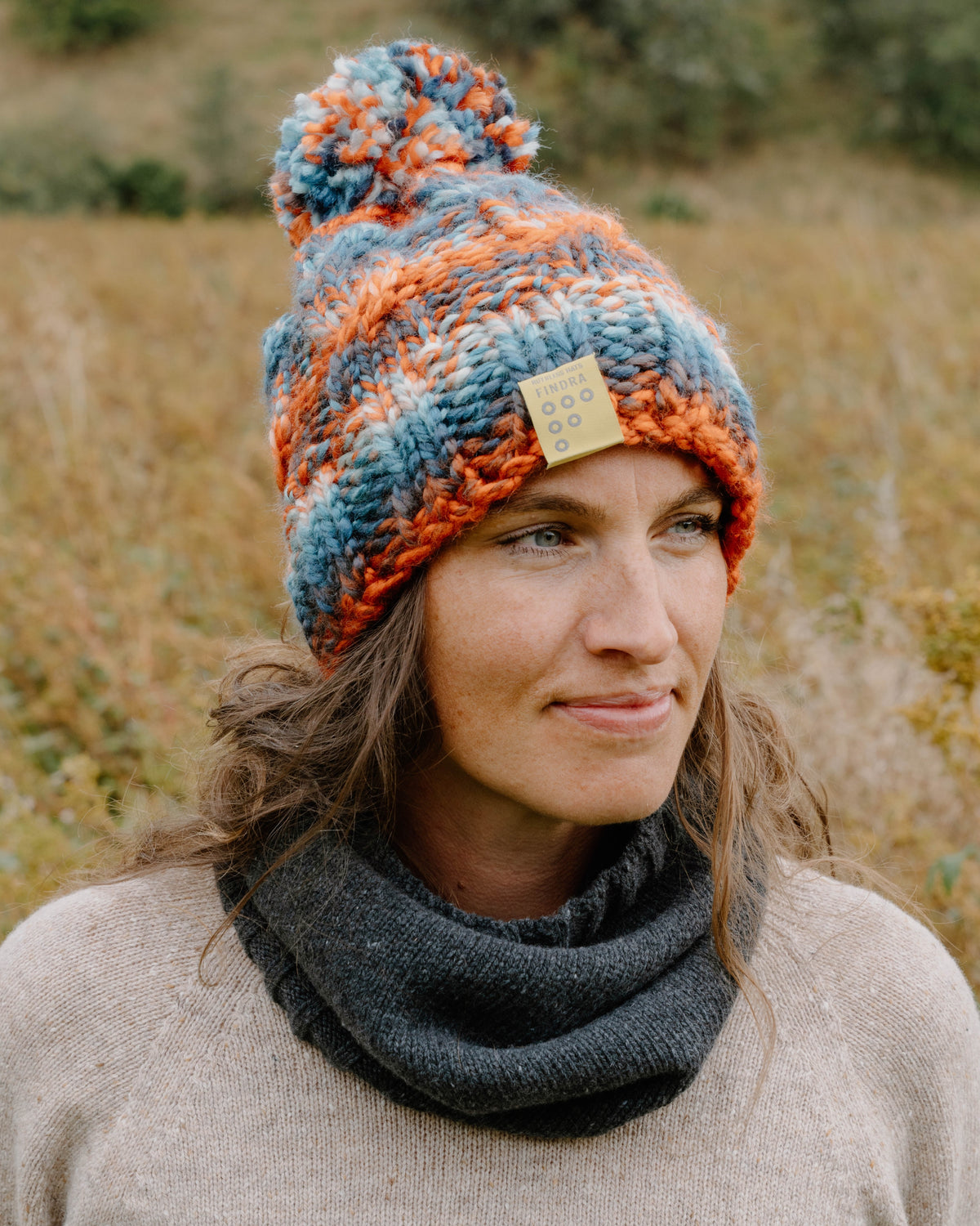 A woman wearing a chunky knit pom-pom bobble hat in blue, orange, and gray, paired with a dark gray cowl neck warmer, photographed outdoors in a field.