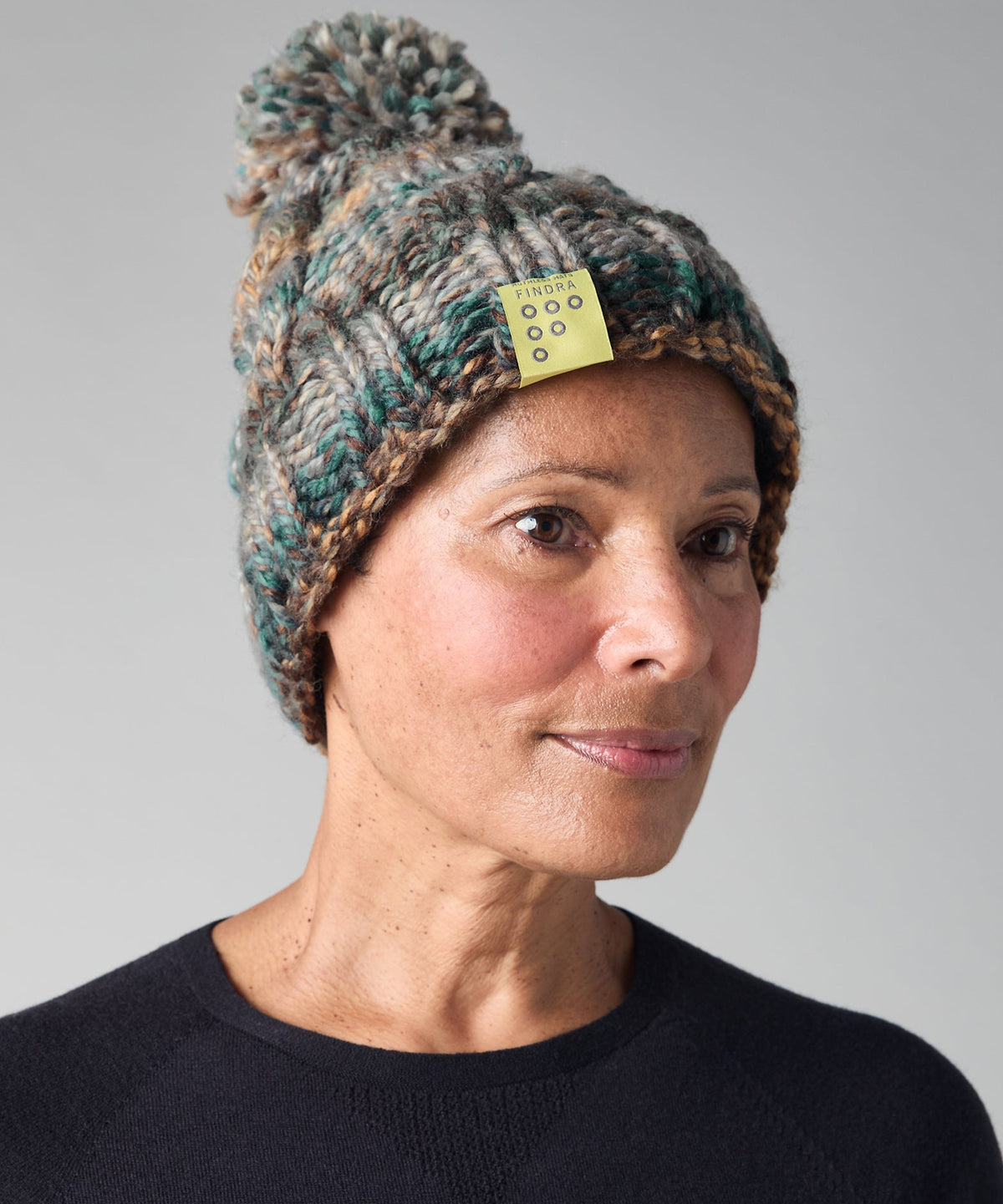 A woman wearing a chunky knit pom-pom beanie in a heathered mix of teal, brown, and cream, with a small yellow tag attached. She is wearing a dark navy crewneck sweater against a neutral gray background.
