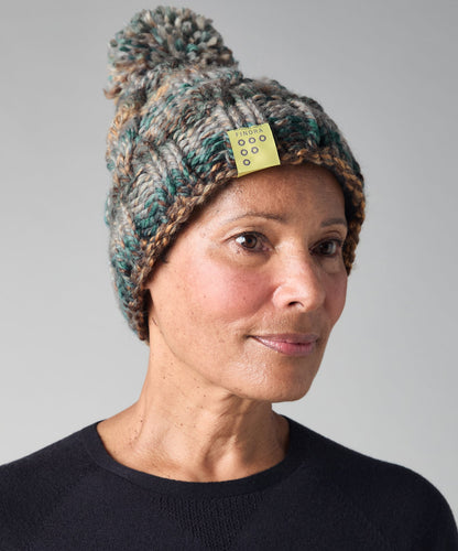 A woman wearing a chunky knit pom-pom beanie in a heathered mix of teal, brown, and cream, with a small yellow tag attached. She is wearing a dark navy crewneck sweater against a neutral gray background.