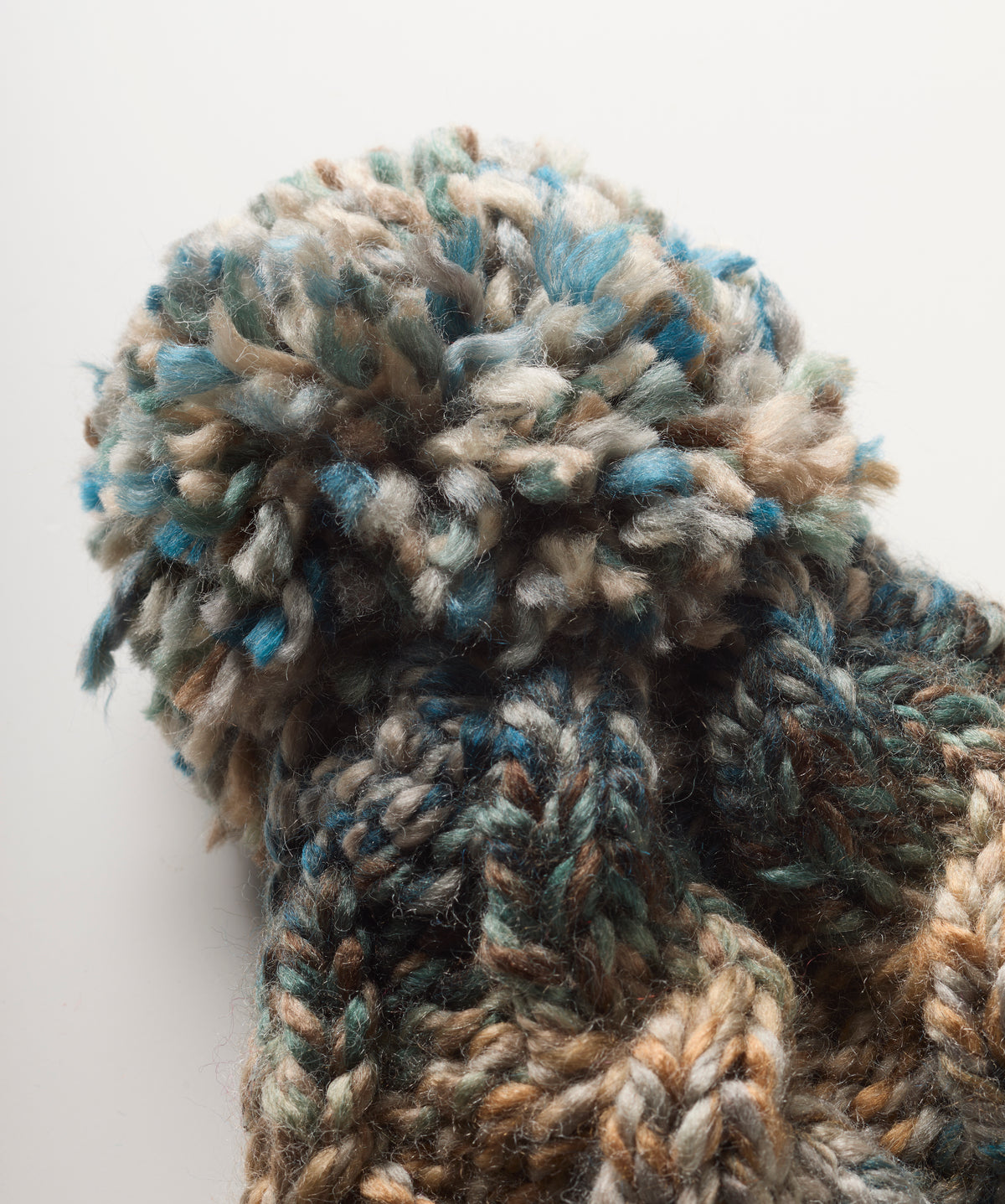 Close-up of a chunky knit bobble hat with a large fluffy pom-pom, made from variegated yarn in blue, beige, brown, and gray tones, set against a plain light background.