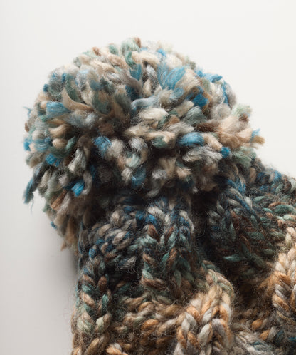 Close-up of a chunky knit bobble hat with a large fluffy pom-pom, made from variegated yarn in blue, beige, brown, and gray tones, set against a plain light background.