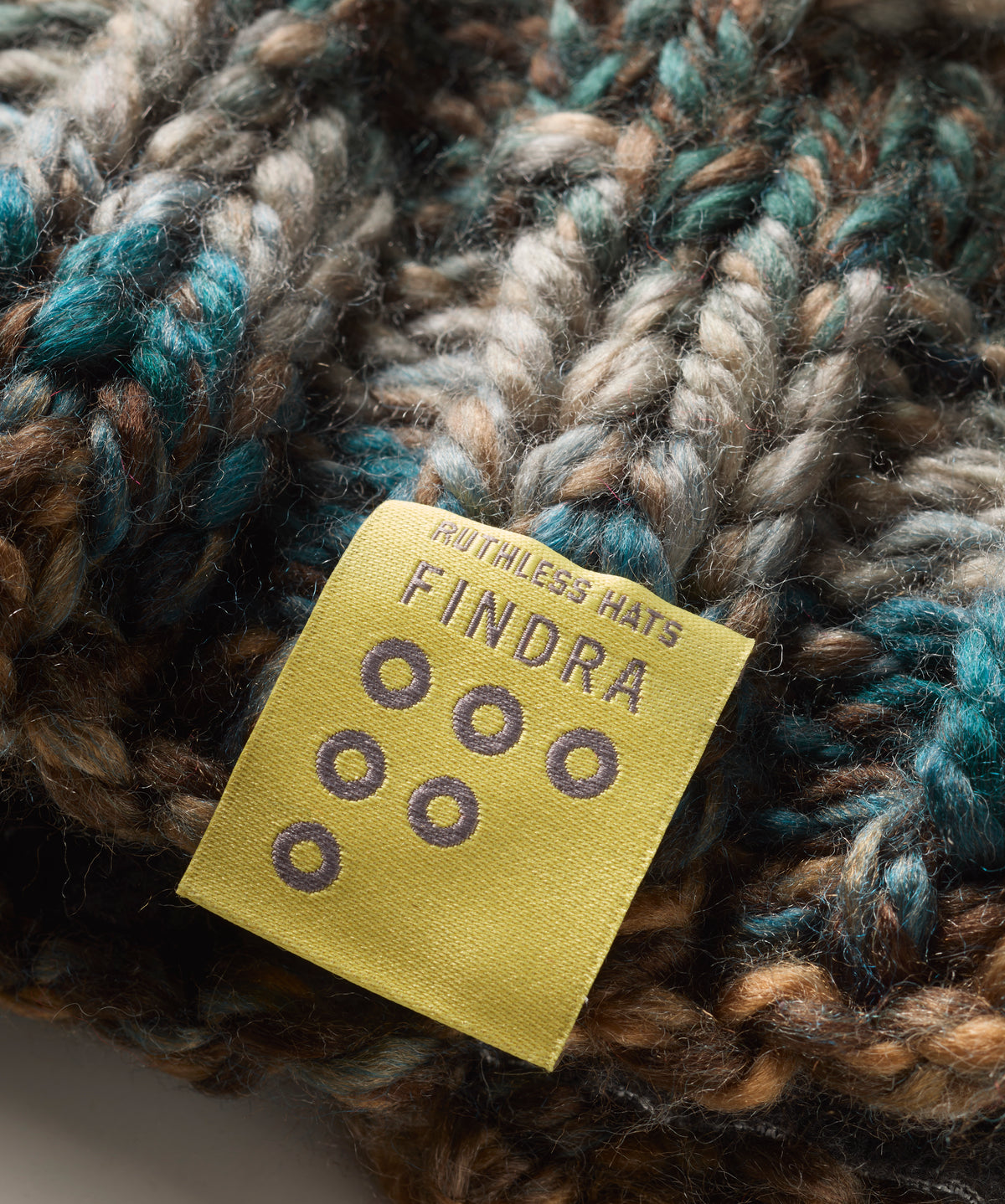 Close-up of a chunky knit hat in mixed brown, beige, and teal yarn with a yellow sewn-in label reading 'FINDRA'.