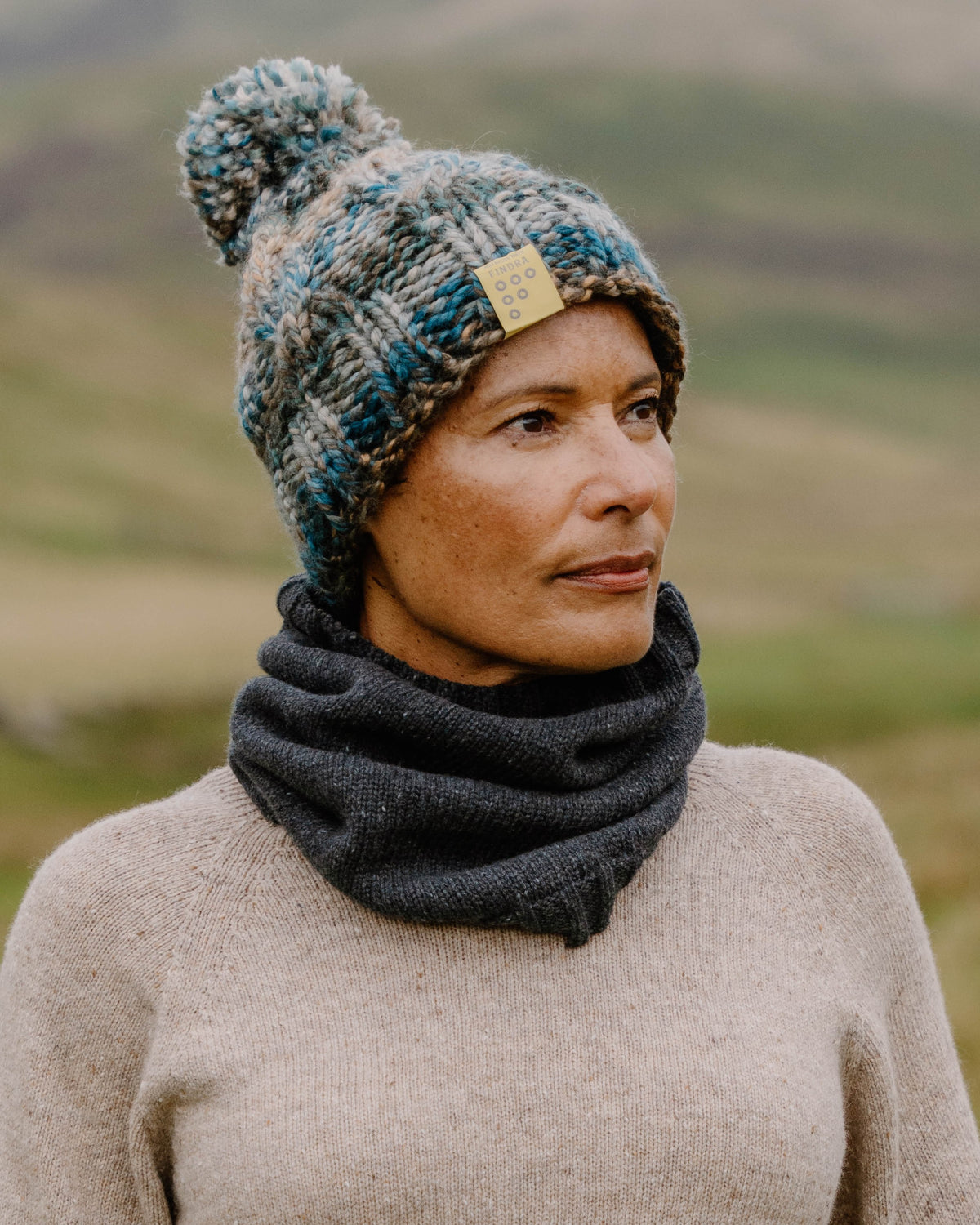 A woman wearing a chunky knit pom-pom beanie in blue, gray, and beige, paired with a dark gray knit neck warmer and a beige sweater, set against a blurred outdoor mountain background.