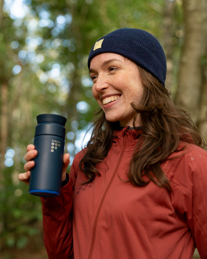 Woman in a red coat and navy beanie in the outdoors enjoying a drink from a Ocean Blue Brew Flask by Ocean bottle and FINDRA