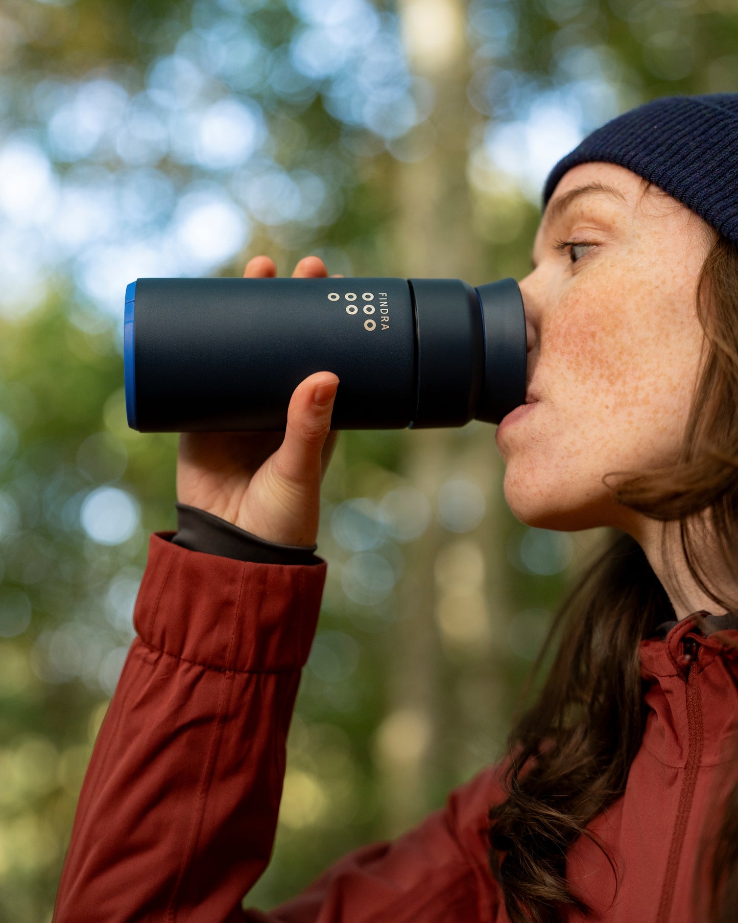 Woman in a red coat and navy beanie in the outdoors enjoying a drink from a Ocean Blue Brew Flask by Ocean bottle and FINDRA
