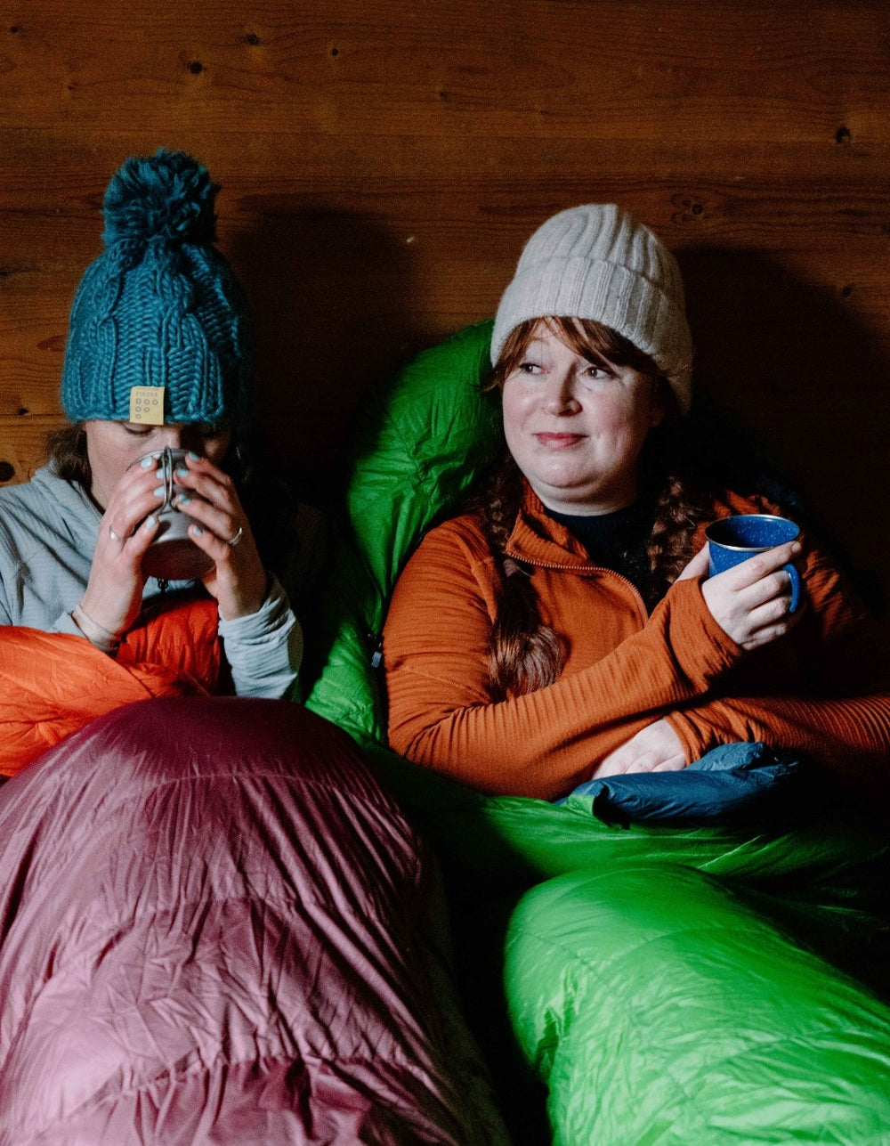 Two people sitting in sleeping bags, one holding a mug, against a wooden wall.
