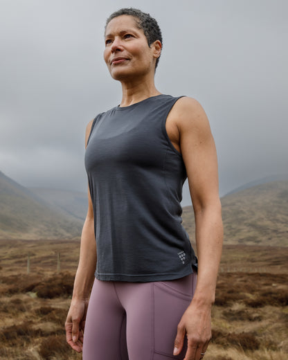 Woman wearing a dark gray sleeveless top and pink leggings standing in a mountainous landscape.