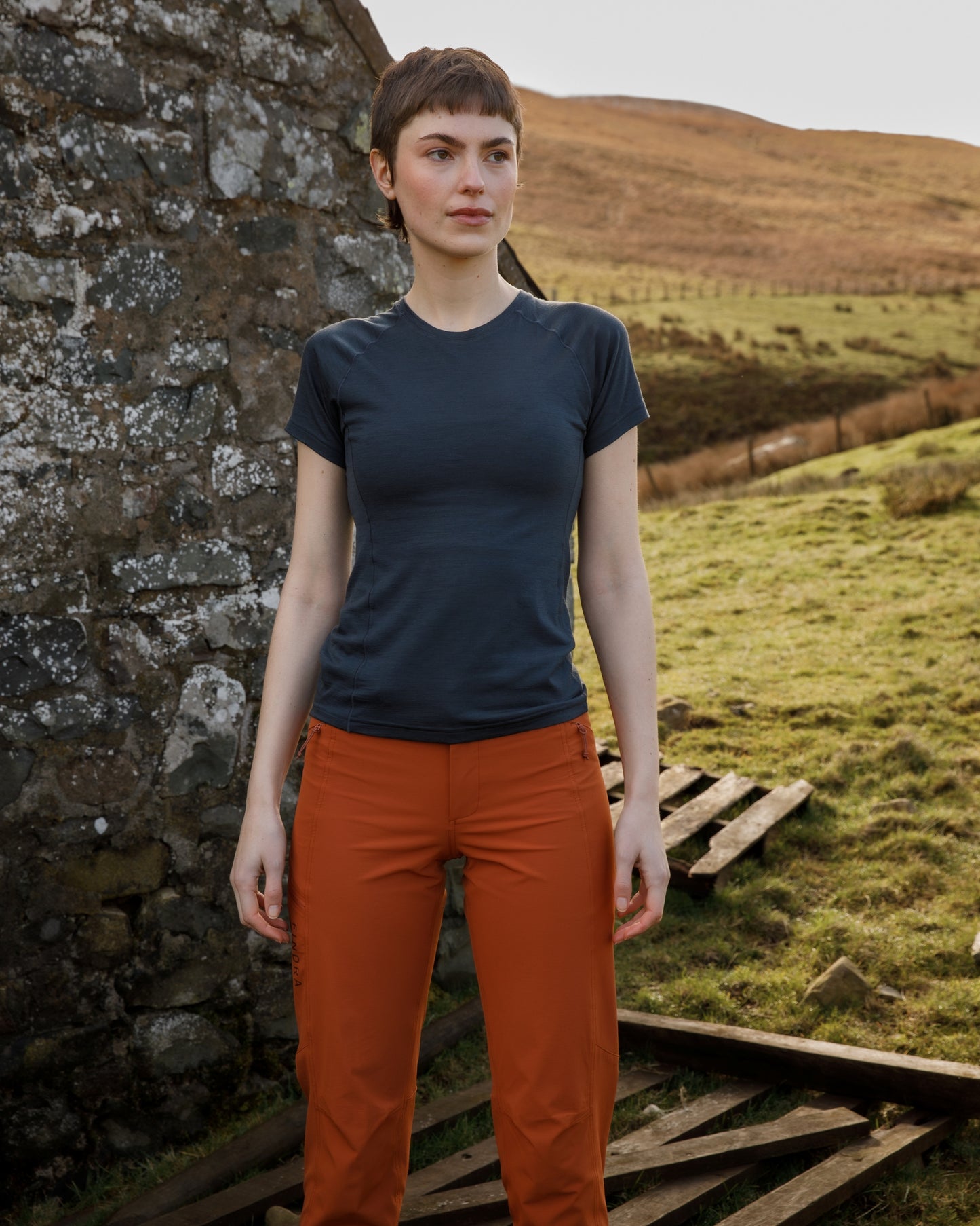 Woman standing in a scenic outdoor setting with a stone wall and grassy field.