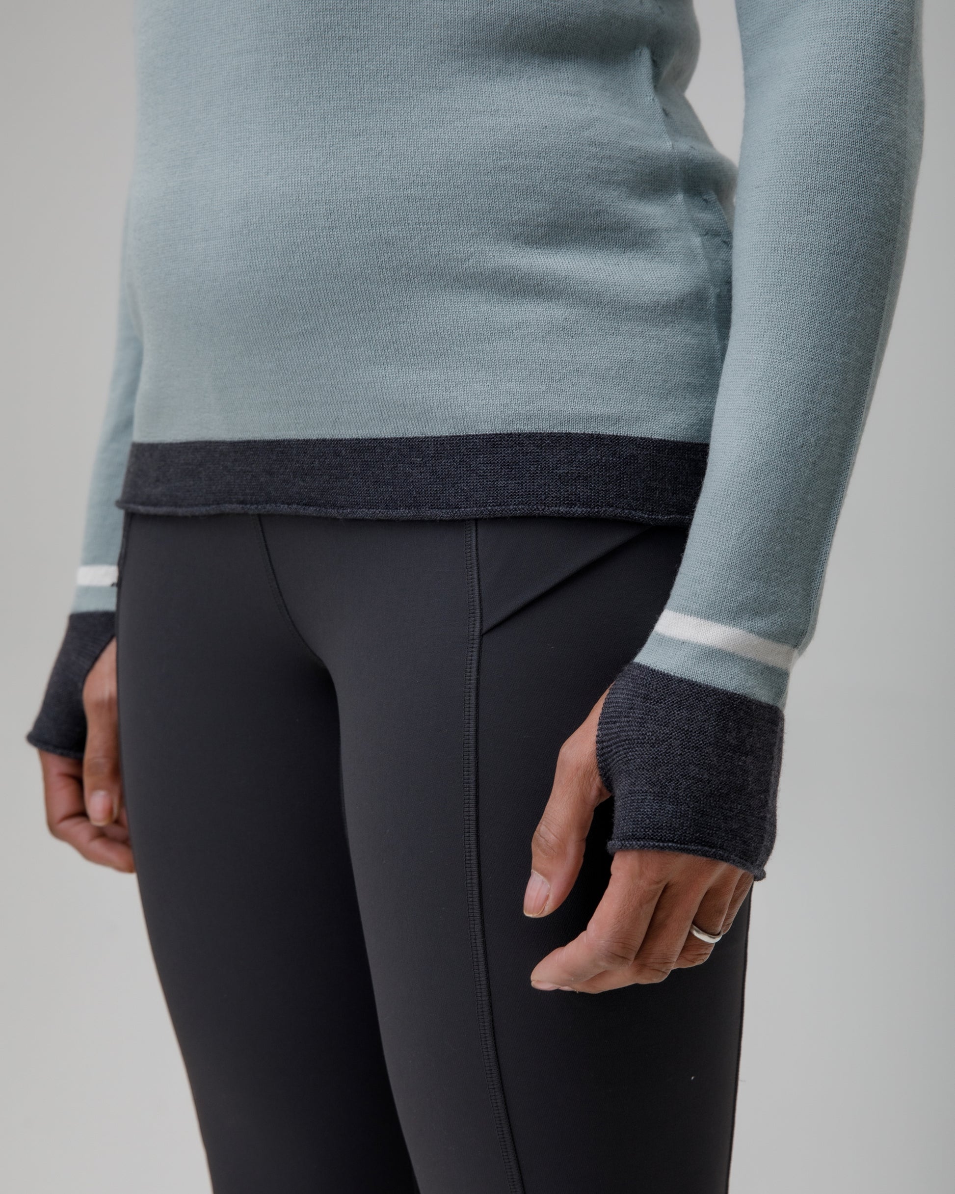 Person wearing a light blue grey Merino top with dark cuffs and white stripes, paired with black pants on a neutral background.
