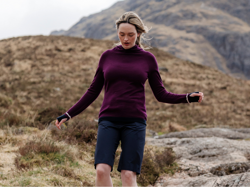 Woman walking down a hillside in a mountainous landscape with barren hills and a cloudy sky in a Merino Wool Hooded top from FINDRA