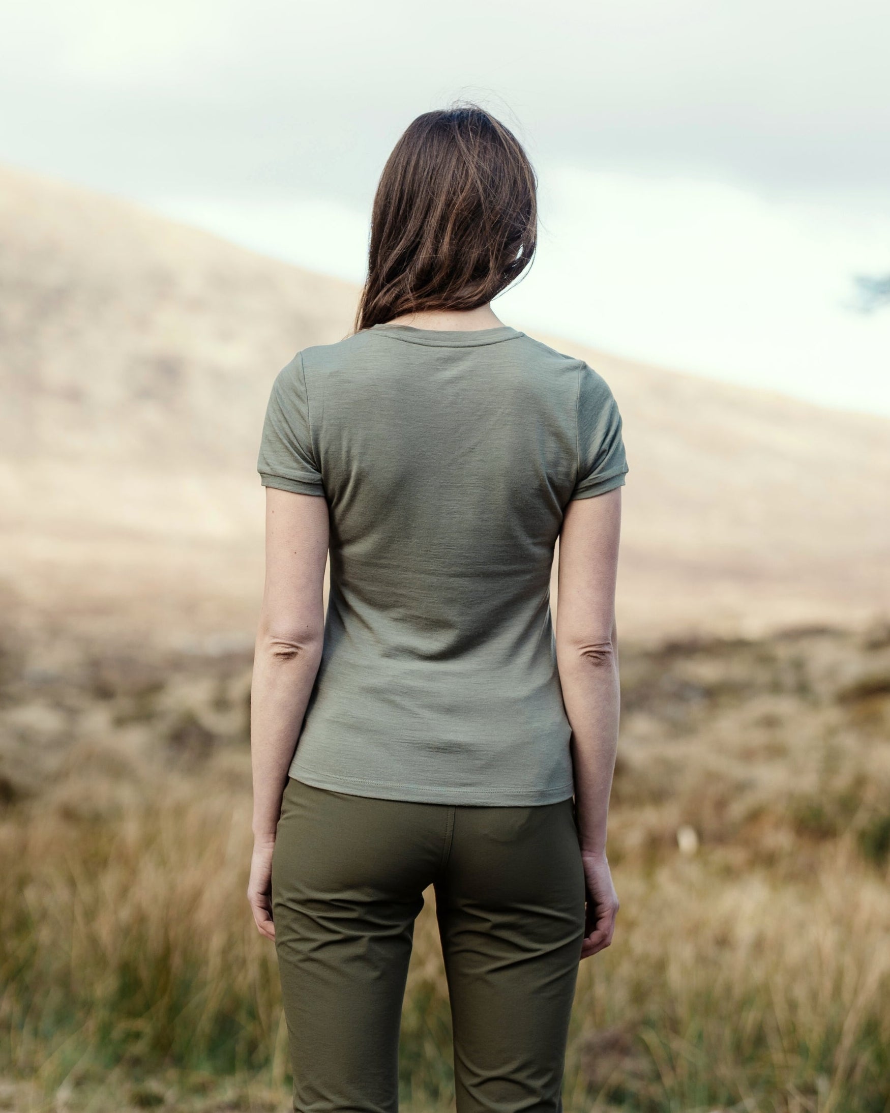 A woman viewed from behind wearing an olive green short-sleeve t-shirt and matching olive green pants, standing in a dry grassy field with a hill in the background.