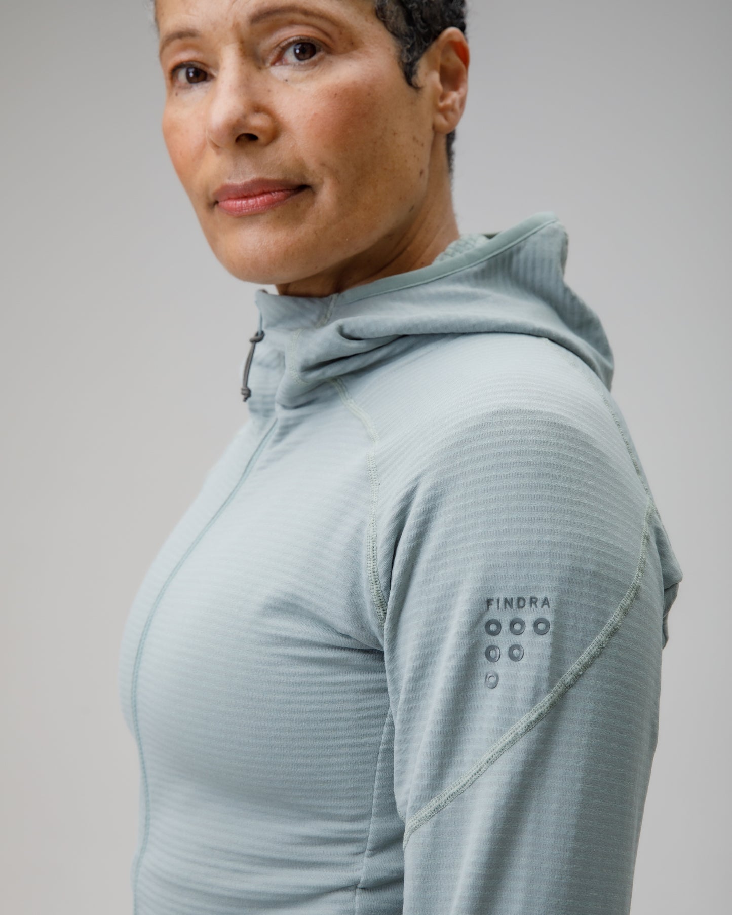 Person wearing a light gray hoodie with brand logo on sleeve against a plain background