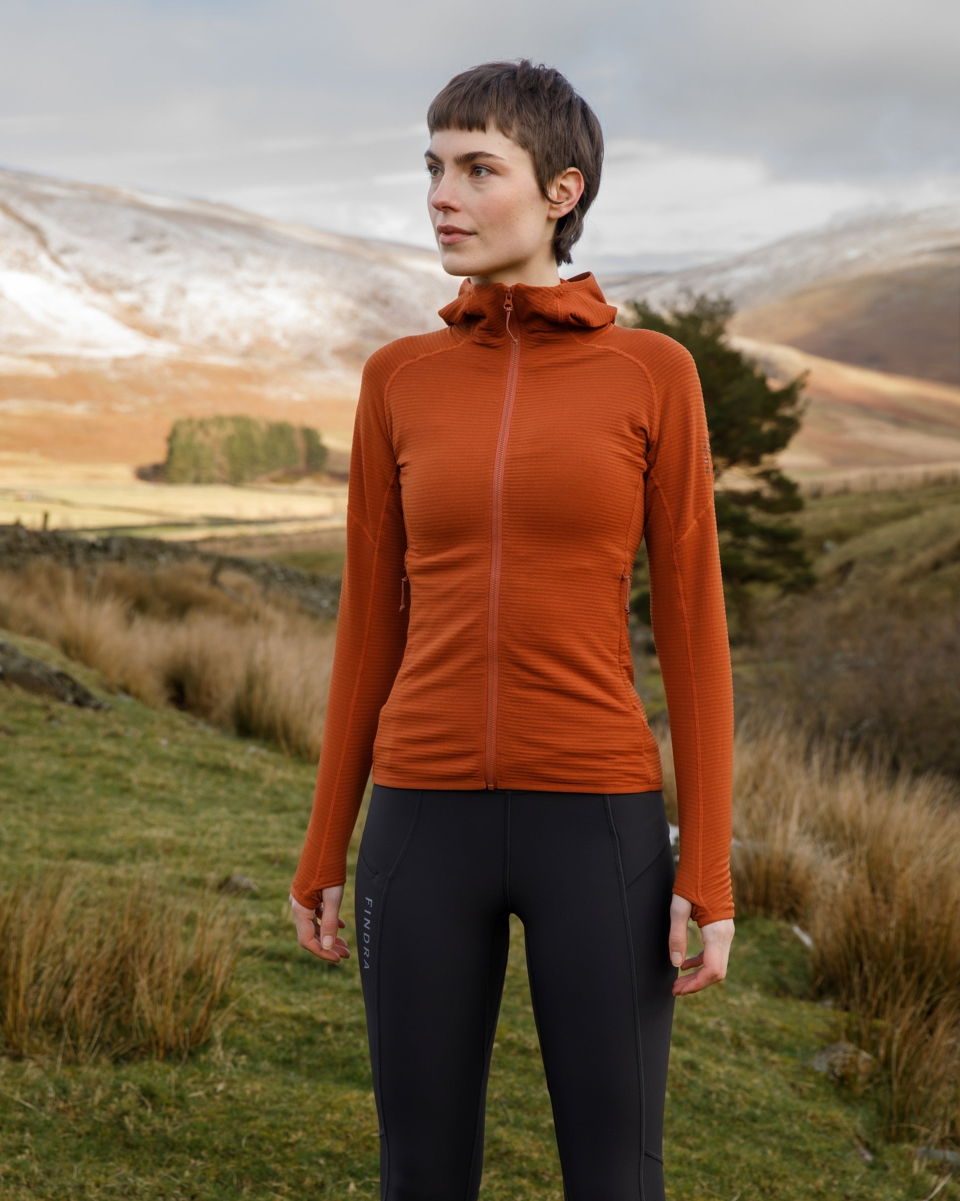 Woman wearing an orange jacket and black pants standing in a scenic outdoor setting with mountains.
