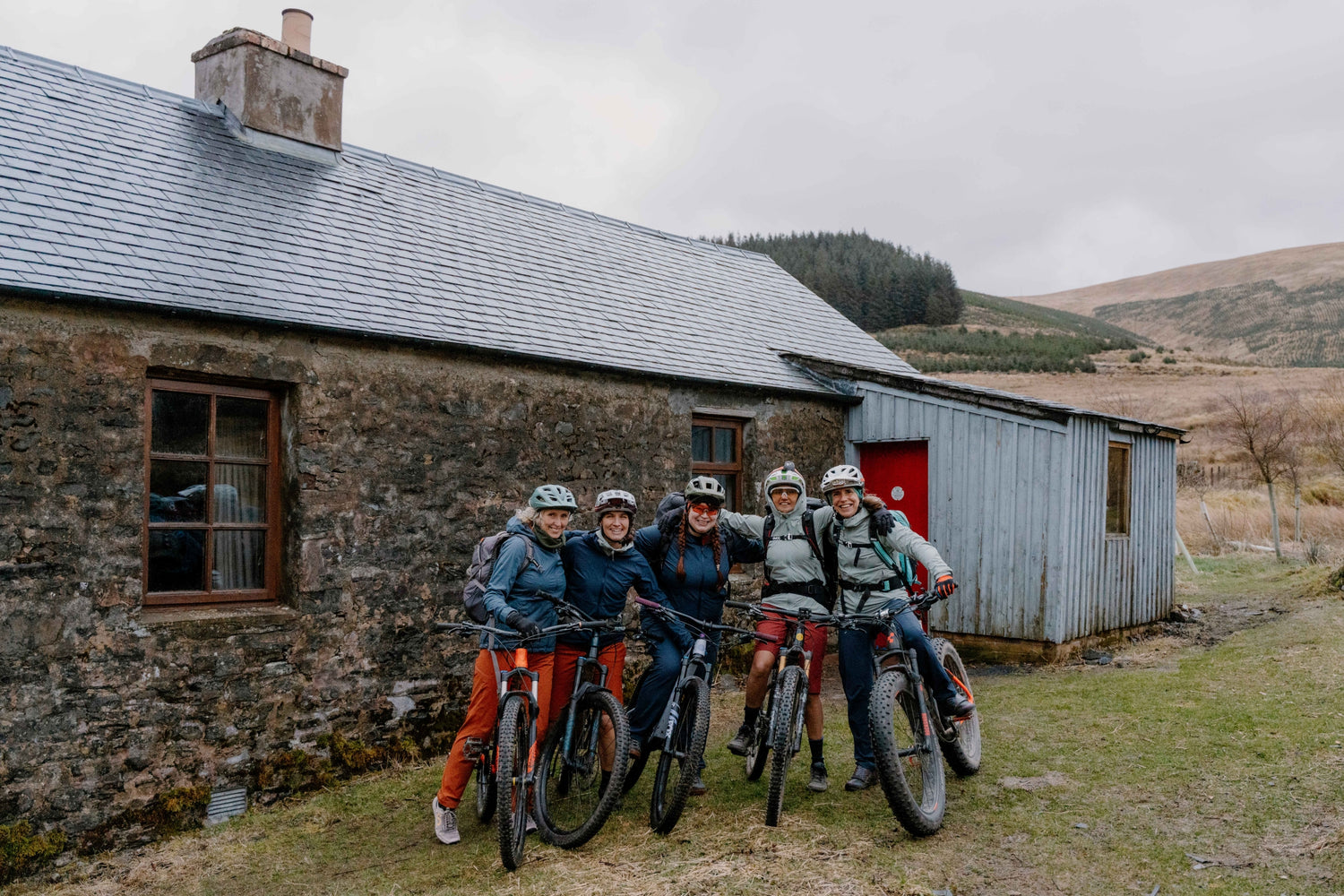 Four people on bicycles in front of a rustic building with a mountainous background