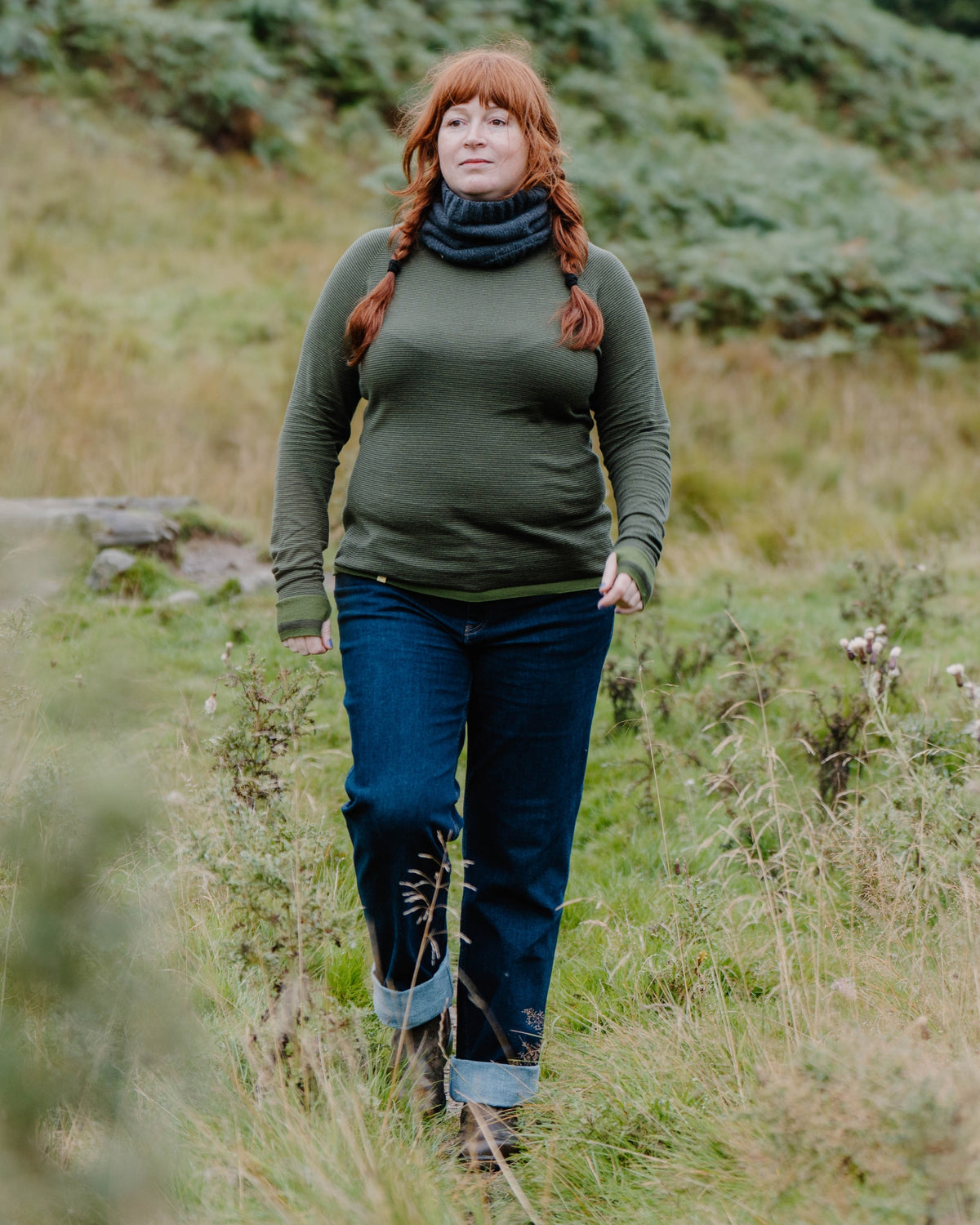 A woman with red hair in braids walks through a grassy field, wearing a Merino wool base layer, dark blue jeans with rolled cuffs, and a dark navy knit cowl scarf.