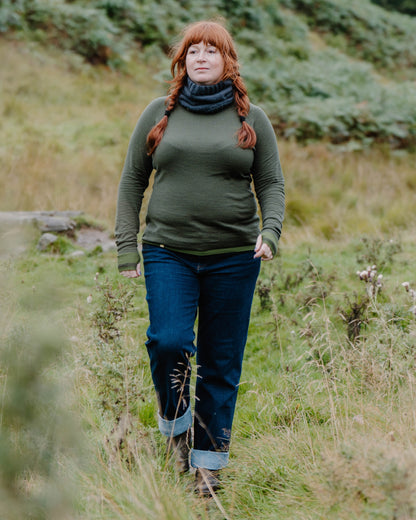 A woman with red hair in braids walks through a grassy field, wearing a Merino wool base layer, dark blue jeans with rolled cuffs, and a dark navy knit cowl scarf.