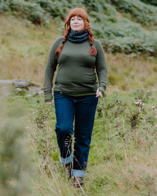 A woman with red hair in braids walks through a grassy field, wearing a Merino wool base layer, dark blue jeans with rolled cuffs, and a dark navy knit cowl scarf.