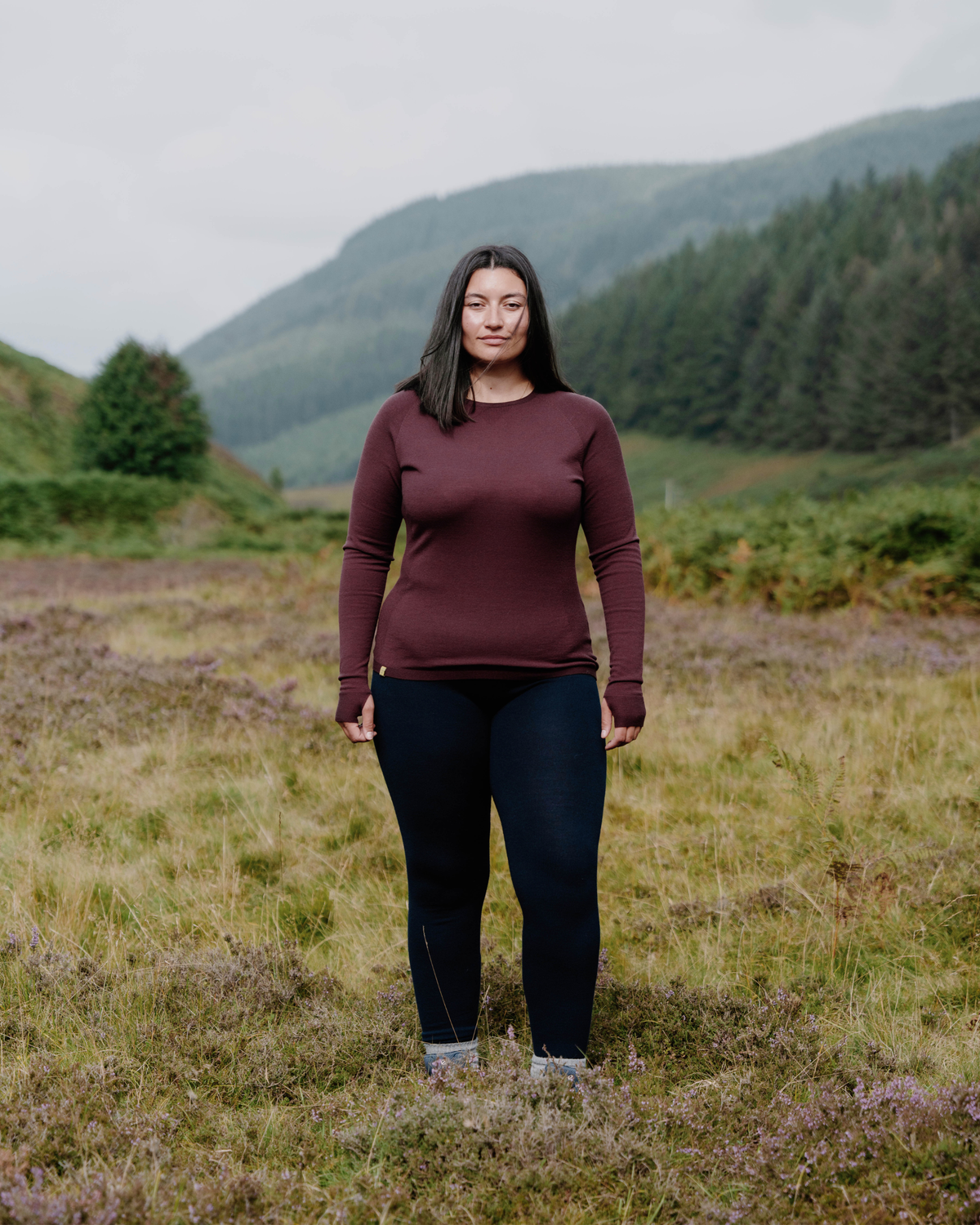 A woman stands in a grassy field with mountains and a forest in the background, wearing a long-sleeve burgundy top and dark navy pants.